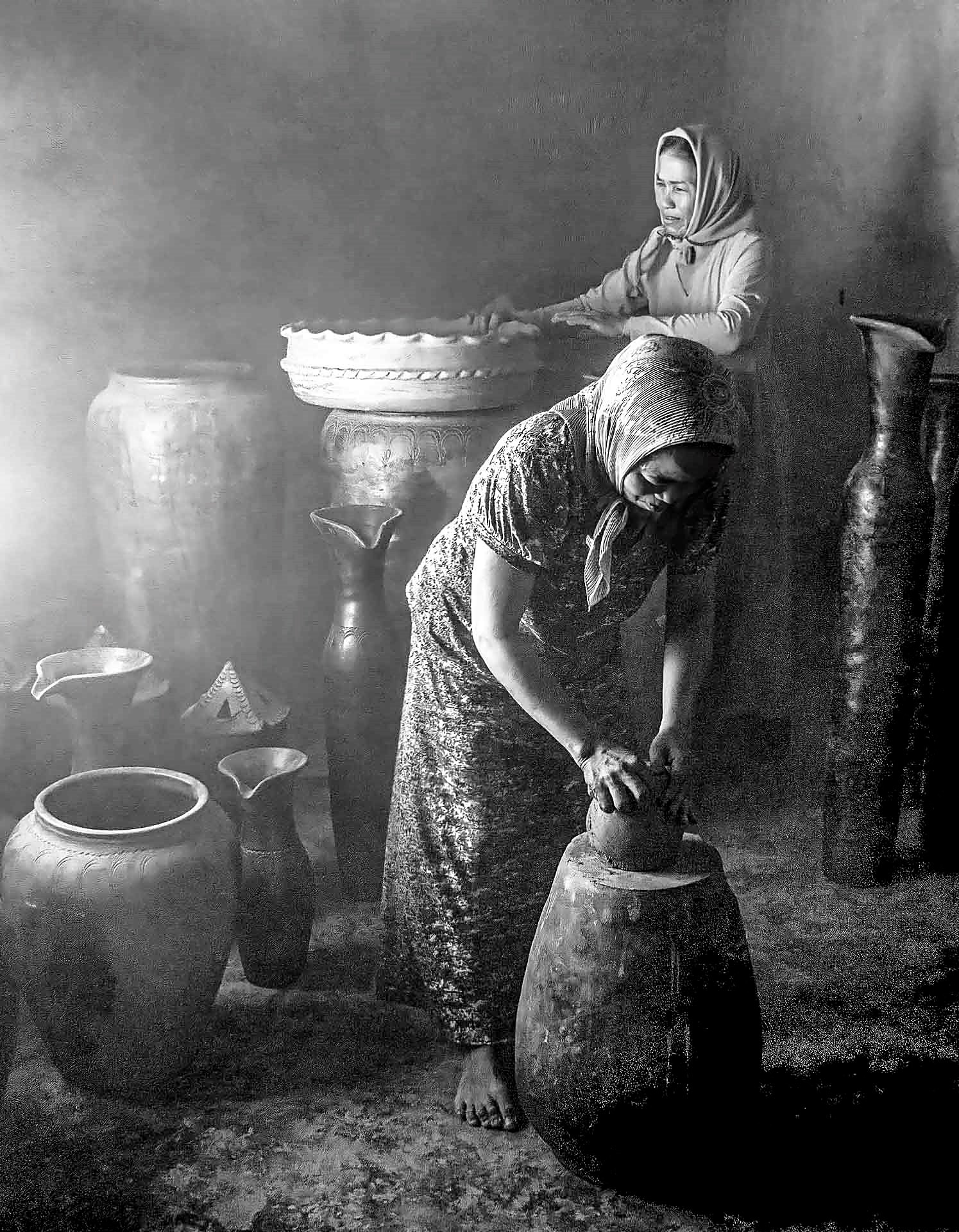 Hai người thợ nữ nặn gốm trong xưởng, quanh là những bình mộc cao