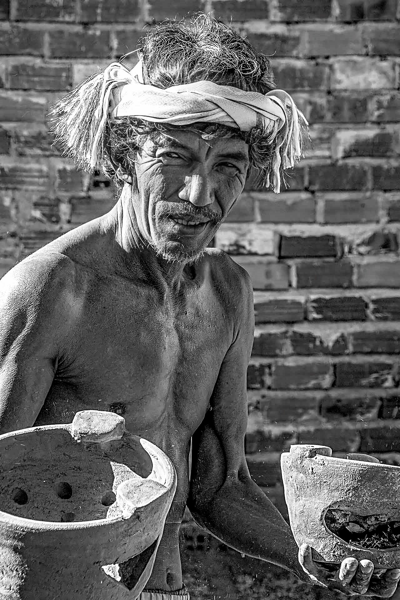 Người thợ nam khăn quấn đầu, hai tay cầm hai món gốm mộc, nền tường gạch